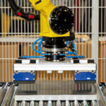 Gelber Industrieroboterarm mit Greifvorrichtung, der ein rechteckiges Metallobjekt auf einer Rollenförderanlage hebt, vor einem weißen Gitterzaun