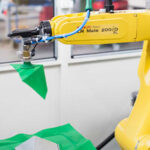 Gelber Industrieroboterarm der Marke FANUC LR Mate 200iD mit Greifer, der ein grünes Tuch hält, in einer Labor- oder Werkstattumgebung