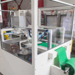 Automatisierte industrielle Maschine mit durchsichtiger Schutzabdeckung, die grüne Kunststoffbahnen verarbeitet, in einer Werkhalle mit Regalen und Werkzeugen im Hintergrund