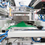 Industrielle Maschine mit Förderband und blauen Kabeln in einer Produktionsanlage, die grüne Platten oder Blätter transportiert