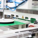 Industrielle Maschine mit Greifarm und Saugteller, die ein grünes Blatt Material auf einem Förderband verarbeitet, umgeben von Kabeln und metallischen Komponenten.