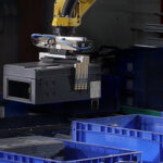 Industrieroboter mit Greifarm, der über mehreren blauen Kunststoffboxen in einer Werkhalle positioniert ist