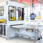 Moderne industrielle Maschine mit mehreren Türen, einem Kontrollpanel mit Bildschirm und einem großen flachen Arbeitstisch in einer hellen Fabrikumgebung