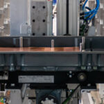 Industrielle Fertigungsanlage mit einem schwarzen Kunststoffbehälter auf einer Maschine, umgeben von Metallrahmen und elektrischen Kabeln in einem Fabrikumfeld.