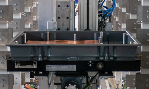Industrielle Fertigungsanlage mit einem schwarzen Kunststoffbehälter auf einer Maschine, umgeben von Metallrahmen und elektrischen Kabeln in einem Fabrikumfeld.
