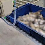 Industrieroboterarm neben einem blauen Behälter mit unscharfem Inhalt in einer Fertigungsanlage.