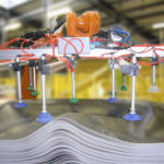 Industrielle Maschine mit mehreren Saugnäpfen und Stangen, die über einem Stapel gewellter, grauer Platten in einer Fabrikumgebung positioniert ist.