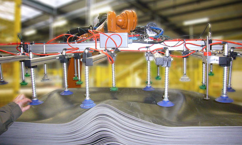 Industrielle Maschine mit mehreren Saugnäpfen und Stangen, die über einem Stapel gewellter, grauer Platten in einer Fabrikumgebung positioniert ist.