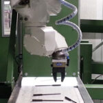 Roboterarm in einer Fertigungsumgebung über einem Förderband mit mehreren Metallstiften darauf