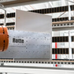 Nahaufnahme eines industriellen Roboters mit einem Metallteil, auf dem "Rotte" steht, vor einem Regal mit nummerierten Fächern in einer hell beleuchteten Werkstatt oder Fabrikumgebung.