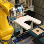 Gelber Industrieroboterarm mit Greifer hält ein weißes, U-förmiges Bauteil in einer automatisierten Fertigungsumgebung mit Zielscheiben auf einem Förderband im Hintergrund.