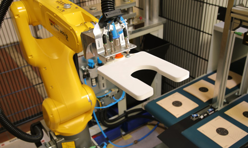 Gelber Industrieroboterarm mit Greifer hält ein weißes, U-förmiges Bauteil in einer automatisierten Fertigungsumgebung mit Zielscheiben auf einem Förderband im Hintergrund.
