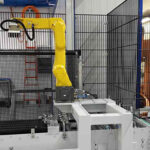 Gelber Industrieroboterarm in geschütztem Bereich einer modernen Fertigungsanlage mit Förderband und Metallumrahmung