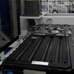 Industrielle Maschine mit Förderband und mehreren schwarzen Plastikbehältern in einer Produktionshalle mit Gitterwand und hellen Wänden im Hintergrund