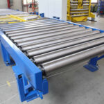 Industrielle Rollbahn aus Metallrollen mit blauer Halterung in einer Werkstattumgebung mit Regalen und Arbeitsbereichen im Hintergrund