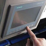 Hand tippt auf ein kleines Siemens-Bedienpanel mit Display und Tasten, das an einer Maschine montiert ist.