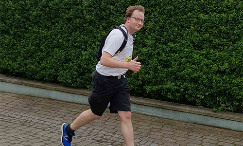 Junger Mann mit Brille, weißem T-Shirt, schwarzer Hose und blau-schwarzen Sportschuhen beim Joggen auf einem gepflasterten Weg vor einer grünen Hecke, der Daumen nach oben zeigend.