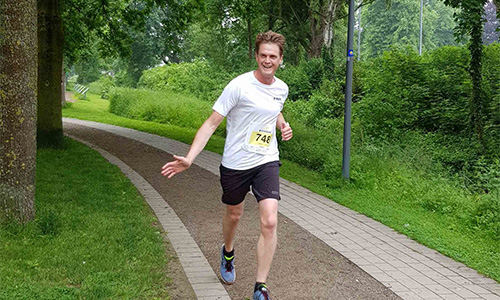 Mann läuft auf einem geschwungenen Weg in einem grünen Park, trägt weißes T-Shirt, schwarze Shorts und Startnummer 748, lächelt und zeigt Daumen hoch.