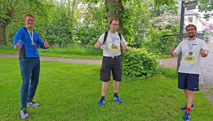 Drei Männer mit Medaillen um den Hals und Startnummern auf der Brust stehen auf einer Wiese neben einem Baum, alle zeigen Daumen nach oben und tragen Sportkleidung.