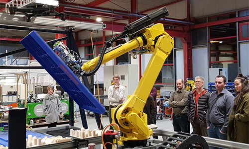 Gelber Industrieroboterarm mit blauem Greifmechanismus, umgeben von mehreren Menschen in einer Werkstatt mit Regalen und Maschinen im Hintergrund.