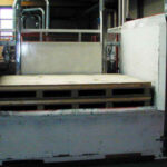 Eine Nahaufnahme einer leeren Holzpalette in einer grauen Industrieumgebung mit Metallgeländern und Maschinen im Hintergrund.