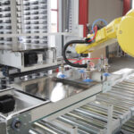 Industrieroboterarm in Gelb, der ein Metallblech auf einem Förderband mit Walzen in einer Fabrikumgebung handhabt.