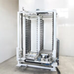 Große weiße industrielle Maschine mit Metallgestell und mehreren vertikalen Reihen von kleinen schwarzen Halterungen in einem hellen, leeren Raum mit Betonboden und weißen Wänden.