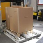 Großer brauner Karton auf einer Palette mit Rollen in einer Lagerhalle oder Fabrikumgebung mit technischen Geräten und Kabeln im Hintergrund