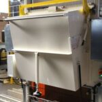 Industriegerät mit geöffnetem Schieber oder Klappe in einer Werkstattumgebung mit Geländer und weiteren Maschinen im Hintergrund