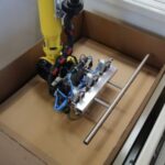 Gelber Industrieroboterarm mit einer montierten Vorrichtung aus Metall und Sensoren, die sich über einer leeren braunen Kartonschachtel befindet.