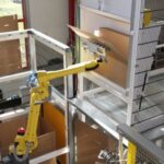 Gelber Industrieroboterarm in einer Fabrikumgebung, der in Richtung eines Computerbildschirms oder Panels zeigt, umgeben von Metallgittern und Geländern.