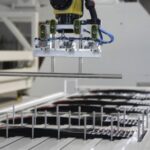 Industrieroboter mit Greifarm, der Metallstäbe über einer Produktionslinie mit mehreren Reihen von eingesteckten Metallstiften hält, in einer Fertigungsumgebung.