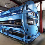 Große blaue Industrieanlage oder Maschine in einer Halle, mit sichtbaren Metallkomponenten und rotem Stahlrahmen im Hintergrund.