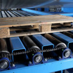 Holzpalette auf einem blauen Förderband mit schwarzen und blauen Walzen und Kettenantrieb in einer Industrieanlage