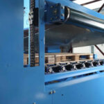 Blau lackierte Industriewerkstattmaschine mit einem horizontalen Fördersystem, Kettenantrieb und mehreren kleinen Rollen auf einer Plattform im Innenraum.