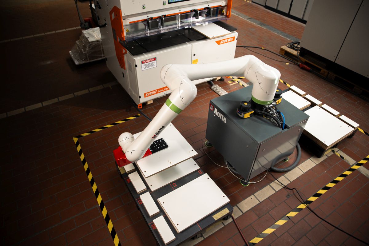Moderner Industrieroboterarm mit weißen Gelenken und grünem Streifen auf einer grauen Basis mit der Aufschrift 'Rotte' in einer Fabrikumgebung mit Maschinen und Holzplatten auf dem Boden