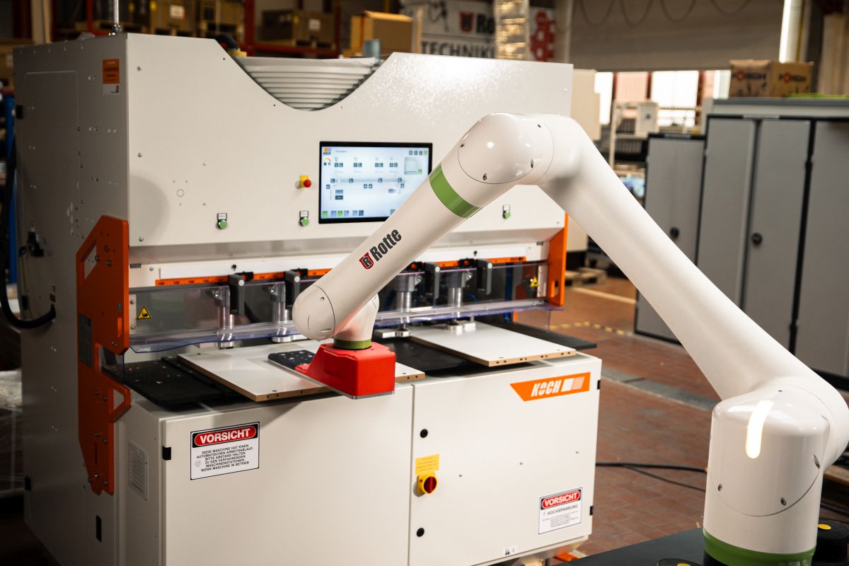 Industrieroboterarm mit dem Markennamen Rotte, der eine Platte vor einer großen weißen und orangefarbenen Maschine mit Monitor in einer Werkhalle bewegt