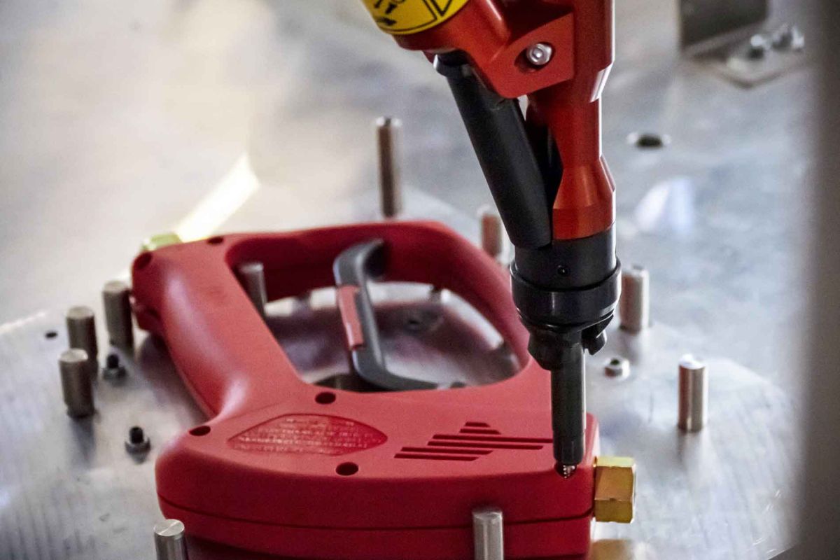 Roboterarm, der an einem roten Kunststoffgriff mit schwarzem Abzug arbeitet, umgeben von Metallstiften auf einer metallischen Oberfläche