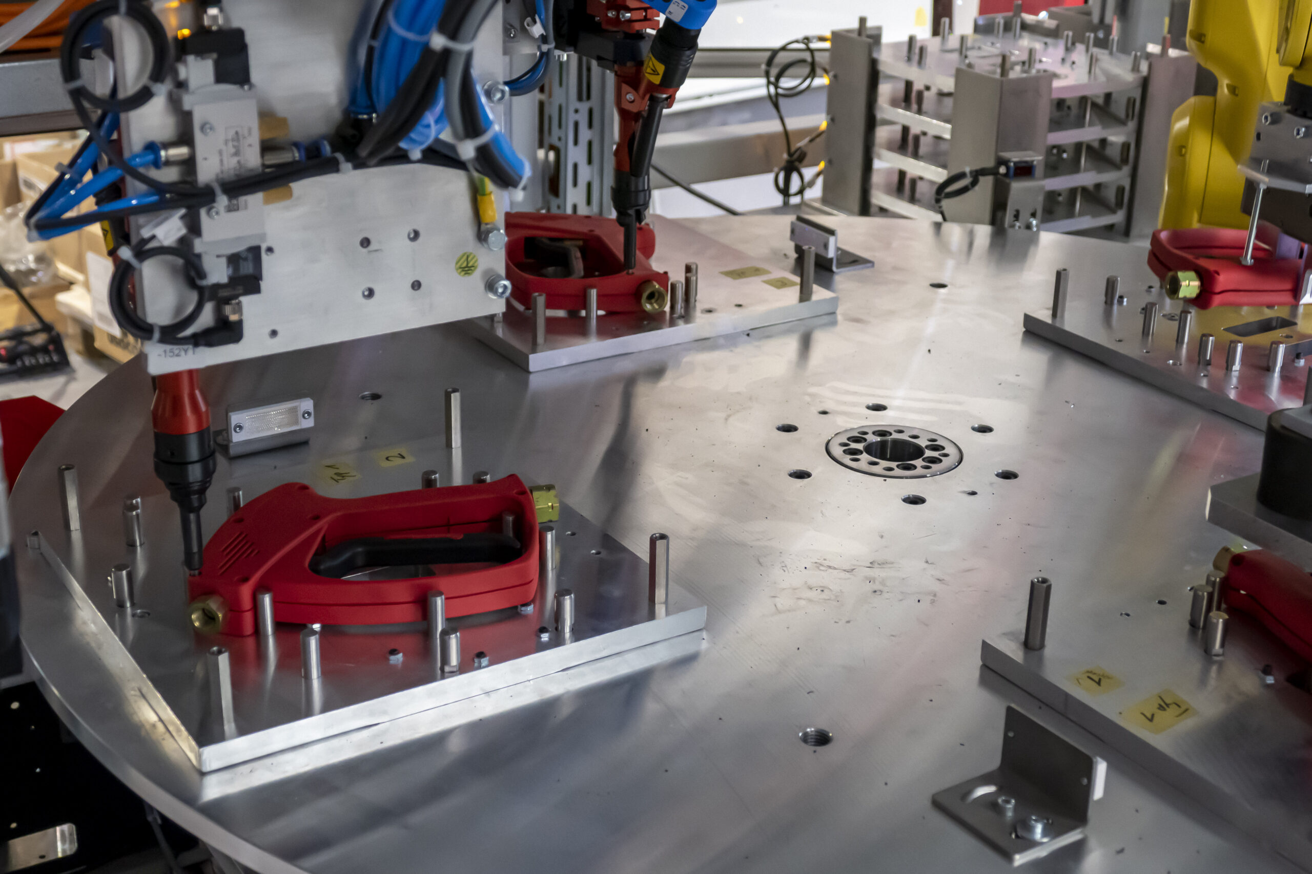 Teil einer industriellen Montagelinie mit rotem Kunststoffgehäuse und automatischen Schraubwerkzeugen auf einer silbernen runden Metallarbeitsfläche.
