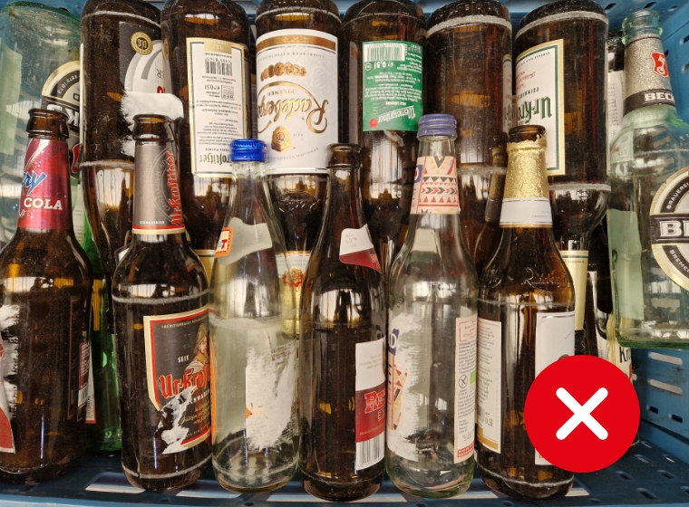 Verschiedene leere und teilweise beschmutzte Bier- und Getränkeflaschen in einer blauen Kiste, vorne ein rotes X-Symbol.