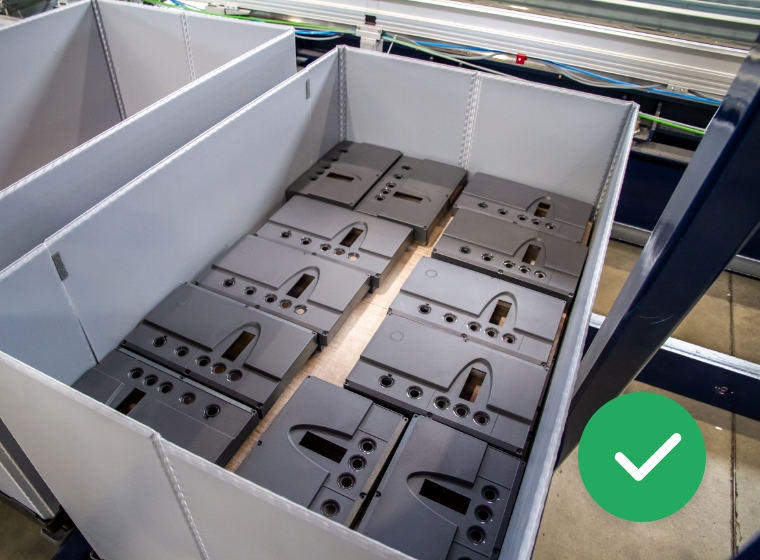 Mehrere gleichförmige graue Metallplatten mit Löchern und Aussparungen liegen ordentlich in einem hellen Kunststoffbehälter auf einem Holzboden in einer Industrieumgebung.