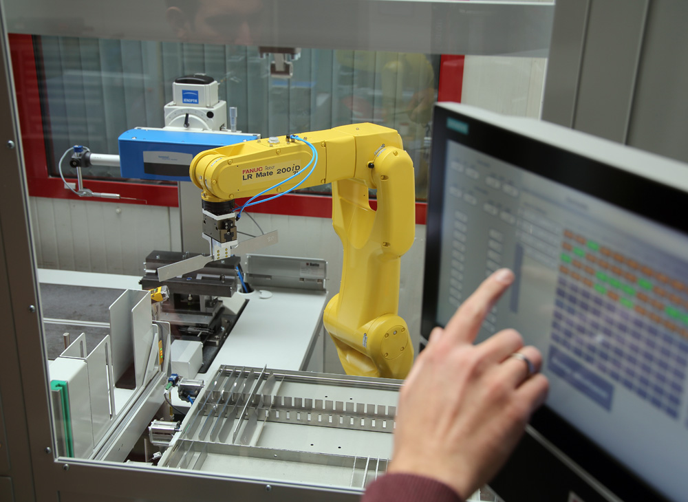 Ein gelber Industrieroboterarm vom Typ FANUC LR Mate 200iD bearbeitet Werkstücke in einer Maschine, während eine Person auf einem Touchscreen Bedienbefehle eingibt.