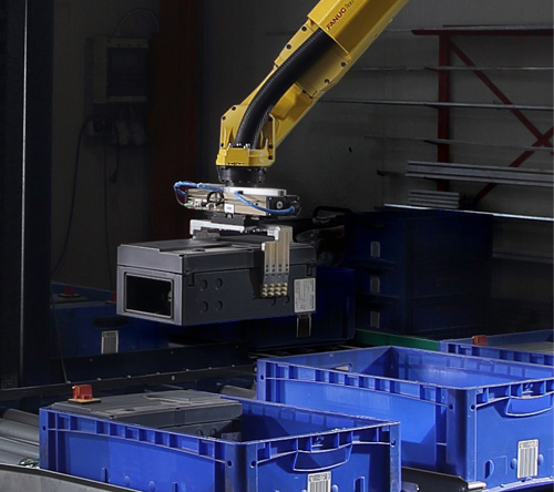 Gelber Industrieroboterarm mit Greifvorrichtung hebt eine schwarze rechteckige Box über mehrere blaue Kunststoffkisten in einer Fabrikumgebung