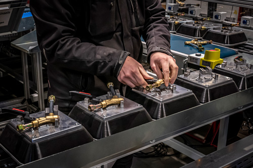 Person in schwarzer Kleidung montiert Metallarmaturen auf einer Produktionslinie mit mehreren identischen Armaturen und maschinellen Vorrichtungen in einer Industrieumgebung.