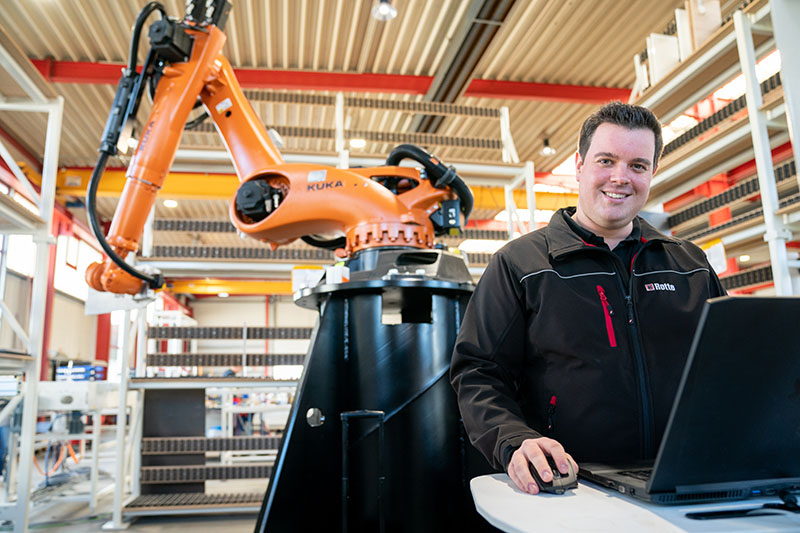 Mann in schwarzer Arbeitsjacke sitzt an einem Laptop in einer Fabrikumgebung mit einem großen orangen Industrieroboterarm der Marke KUKA im Hintergrund.