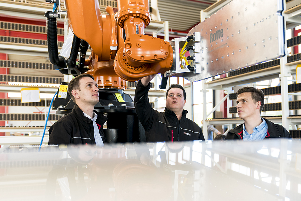 Drei Männer in Arbeitskleidung beobachten und bedienen einen großen orangen Industrieroboterarm in einer Werkhalle mit Regalen im Hintergrund.