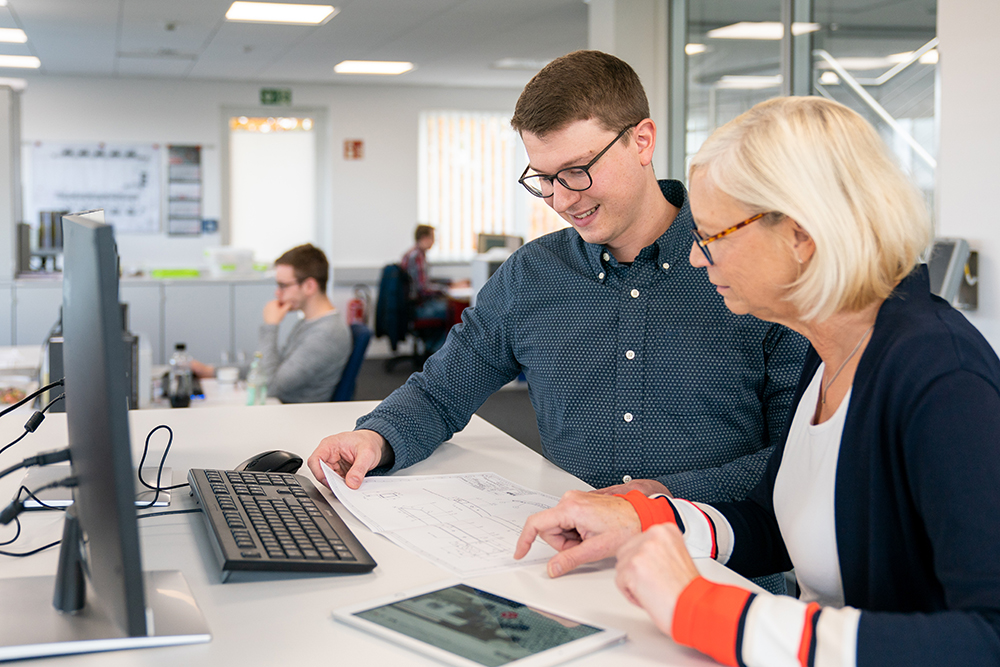 Zwei Kollegen, ein Mann und eine Frau, die zusammen an einem Schreibtisch in einem Büro sitzen, der Mann hält einen Plan auf Papier, die Frau zeigt auf ein Tablet, im Hintergrund weitere Personen an Schreibtischen.