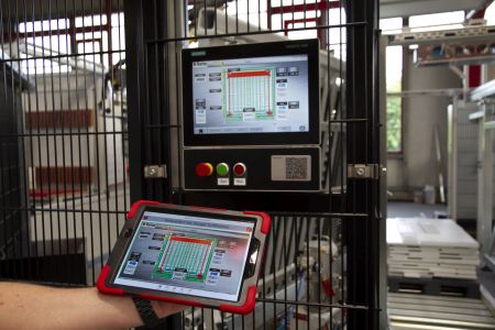 Industriegerät mit Bedienfeld und Monitor hinter Gittern, im Vordergrund eine Hand, die ein Tablet mit einer ähnlichen Benutzeroberfläche hält, in einer Fabrikumgebung.