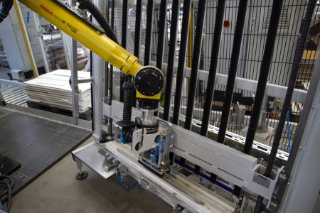 Gelber Industrieroboterarm mit Greifer an einer Produktionsmaschine zum automatischen Handling von weißen Platten in einer Fabrikumgebung