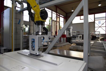 Gelber Industrieroboterarm in einer Fabrikumgebung, der auf einer weißen Oberfläche arbeitet, umgeben von Maschinen und Metallstrukturen
